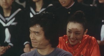 Movie still from “School in the Crosshairs” (1981), directed by Nobuhiko Ôbayashi – A man and a woman sitting next to each other in front of other people; Close Up shot, Over the shoulder angle
