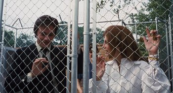 Movie still from “Scorpio” (1973), directed by Michael Winner – A man and a woman standing next to each other in front of a chain link fence; Medium shot, Over the shoulder angle