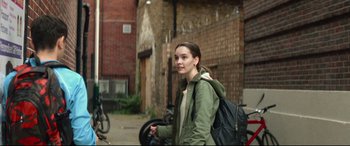 Movie still from “Scrapper” (2023), directed by Charlotte Regan – A woman in a green jacket is walking down the street; Medium shot, Over the shoulder angle