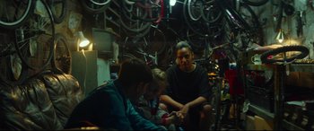 Movie still from “Scrapper” (2023), directed by Charlotte Regan – A group of people sitting in a room next to bicycles; Wide shot, Over the shoulder angle