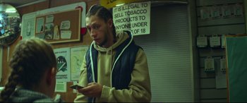 Movie still from “Scrapper” (2023), directed by Charlotte Regan – A man holding a cell phone while standing in a room; Medium shot, Low angle