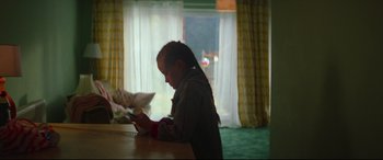 Movie still from “Scrapper” (2023), directed by Charlotte Regan – A young girl sitting at a table looking at her cell phone; Medium shot, Over the shoulder angle