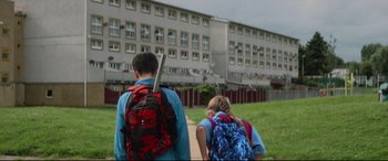 Movie still from “Scrapper” (2023), directed by Charlotte Regan – Two people walking in front of a building; Wide shot, Low angle