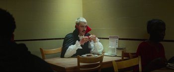 Movie still from “Scrapper” (2023), directed by Charlotte Regan – A man sitting at a table with a plate in front of him; Medium shot, Over the shoulder angle
