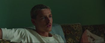 Movie still from “Scrapper” (2023), directed by Charlotte Regan – A man sitting on top of a couch in front of a window; Close Up shot, Over the shoulder angle