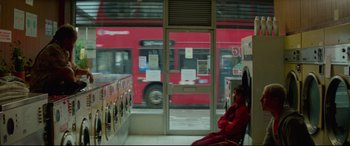 Movie still from “Scrapper” (2023), directed by Charlotte Regan – A person sitting on a bench in front of a bus; Wide shot, Low angle