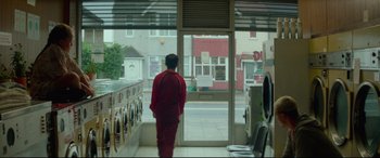 Movie still from “Scrapper” (2023), directed by Charlotte Regan – A man in a red suit looking out of a store window; Wide shot, Low angle