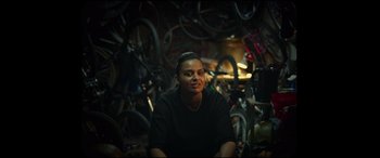 Movie still from “Scrapper” (2023), directed by Charlotte Regan – A man sitting in a garage surrounded by bicycles; Medium shot, Low angle