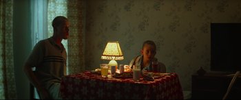 Movie still from “Scrapper” (2023), directed by Charlotte Regan – A child sitting at a table with a lit lamp on it; Medium shot, High angle
