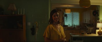 Movie still from “Scrapper” (2023), directed by Charlotte Regan – A woman standing in a kitchen with a smile on her face; Medium shot, Over the shoulder angle