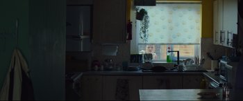 Movie still from “Scrapper” (2023), directed by Charlotte Regan – A young child looking out of a window in a kitchen; Wide shot, Over the shoulder angle