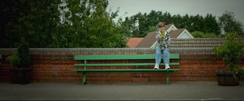 Movie still from “Scrapper” (2023), directed by Charlotte Regan – A boy sitting on top of a green park bench; Wide shot, Low angle