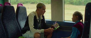 Movie still from “Scrapper” (2023), directed by Charlotte Regan – A man sitting in front of a window holding a cake; Medium shot, Over the shoulder angle