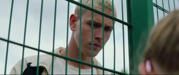 Movie still from “Scrapper” (2023), directed by Charlotte Regan – A man with blonde hair is behind a green fence; Close Up shot, Low angle