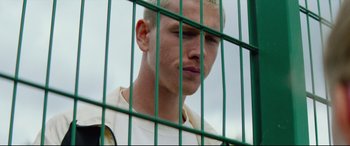 Movie still from “Scrapper” (2023), directed by Charlotte Regan – A man with blonde hair is behind bars; Close Up shot, Low angle