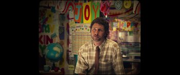 Movie still from “Scrapper” (2023), directed by Charlotte Regan – A man sitting in front of a wall with a lot of different decorations; Medium shot, Over the shoulder angle