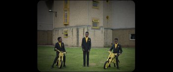 Movie still from “Scrapper” (2023), directed by Charlotte Regan – Three young men in suits and ties standing in front of a building; Extreme Wide shot, High angle