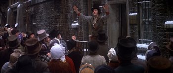 Movie still from “Scrooge” (1970), directed by Ronald Neame – A man in a hat is on a stage with a bunch of people; Wide shot, Low angle