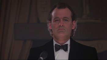 Movie still from “Scrooged” (1988), directed by Richard Donner – A man in a suit and bow tie is standing in front of a microphone; Close Up shot, Low angle