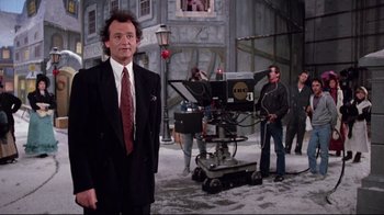 Movie still from “Scrooged” (1988), directed by Richard Donner – A man in a suit and tie standing in front of a camera; Medium shot, Over the shoulder angle