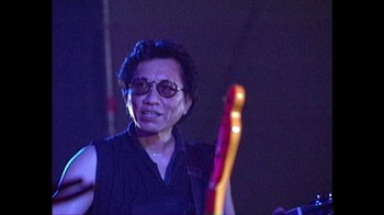 Movie still from “Searching for Sugar Man” (2012), directed by Malik Bendjelloul – An older man holding a guitar in front of a crowd of onlookers; Close Up shot, Low angle