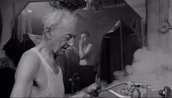 Movie still from “Seconds” (1966), directed by John Frankenheimer – An old man looking in the mirror in front of a man smoking; Medium shot, Low angle