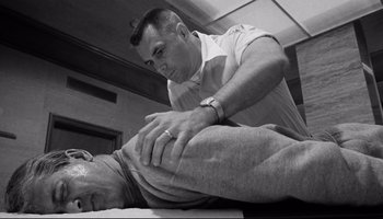 Movie still from “Seconds” (1966), directed by John Frankenheimer – A man in a white shirt is stretching a man's leg; Medium shot, Low angle