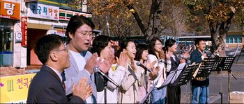 Movie still from “Secret Sunshine” (2007), directed by Lee Chang-dong – A group of people standing next to each other holding microphones; Medium shot, High angle
