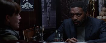 Movie still from “Secret in Their Eyes” (2015), directed by Billy Ray – A man sitting at a table in front of a baseball picture; Medium shot, Over the shoulder angle