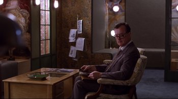 Movie still from “Secretary” (2002), directed by Steven Shainberg – A man sitting in a chair at a desk; Medium shot, Over the shoulder angle
