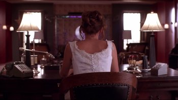 Movie still from “Secretary” (2002), directed by Steven Shainberg – A woman in a white dress sitting at a table in a room; Medium shot, Over the shoulder angle