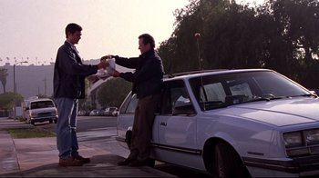 Movie still from “Secretary” (2002), directed by Steven Shainberg – A man handing something to another man standing next to a parked car; Wide shot, Low angle