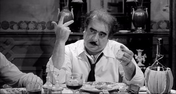 Movie still from “Seduced and Abandoned” (1964), directed by Pietro Germi – An older man sitting at a dinner table holding a piece of cake; Medium shot, High angle