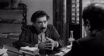 Movie still from “Seduced and Abandoned” (1964), directed by Pietro Germi – A man sitting at a table in front of a window; Medium shot, Over the shoulder angle