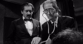 Movie still from “Seduced and Abandoned” (1964), directed by Pietro Germi – Two men in suits and ties with a stethoscope around their neck; Medium shot, Low angle