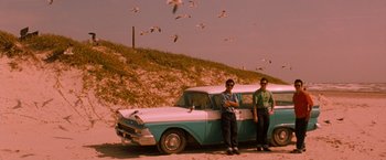 Movie still from “Selena” (1997), directed by Gregory Nava – Two men standing next to an old car on the beach; Wide shot, Low angle