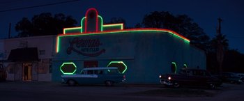Movie still from “Selena” (1997), directed by Gregory Nava – An old car parked in front of a building with neon lights on it; Extreme Wide shot, Low angle