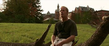 Movie still from “Session 9” (2001), directed by Brad Anderson – A man sitting on a stump in front of a building; Medium shot, Low angle