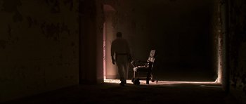 Movie still from “Session 9” (2001), directed by Brad Anderson – A man standing next to a chair in a dark room; Wide shot, Low angle