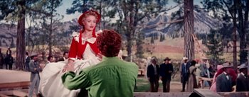 Movie still from “Seven Brides for Seven Brothers” (1954), directed by Stanley Donen – A woman in a white dress sitting on a bench; Medium shot, Over the shoulder angle