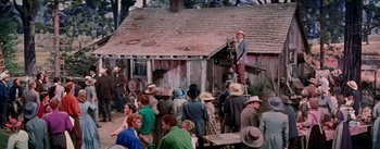 Movie still from “Seven Brides for Seven Brothers” (1954), directed by Stanley Donen – A group of people standing around a wooden house; Wide shot, High angle