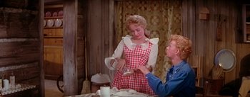 Movie still from “Seven Brides for Seven Brothers” (1954), directed by Stanley Donen – A woman in a checkered dress pouring a glass of milk; Medium shot, Over the shoulder angle