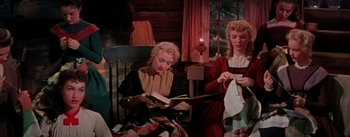Movie still from “Seven Brides for Seven Brothers” (1954), directed by Stanley Donen – Two women are sitting in a chair and one is reading a book while the other one is holding a towel; Medium shot, High angle