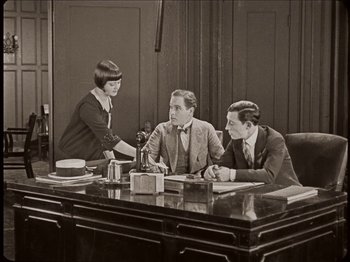 Movie still from “Seven Chances” (1925), directed by Buster Keaton – Three people are sitting at a desk in a room; Medium shot, High angle
