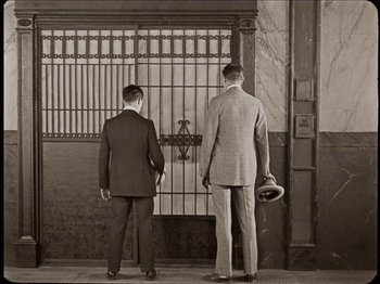 Movie still from “Seven Chances” (1925), directed by Buster Keaton – A couple of men standing next to each other near a gate; Wide shot, Low angle