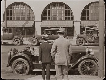 Movie still from “Seven Chances” (1925), directed by Buster Keaton – Two men standing in front of a row of antique cars; Extreme Wide shot, Low angle
