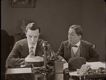 Movie still from “Seven Chances” (1925), directed by Buster Keaton – Two men sitting at a table in front of a phone; Medium shot, High angle