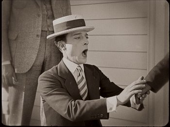 Movie still from “Seven Chances” (1925), directed by Buster Keaton – A man in a suit and a hat is holding his hands up; Medium shot, High angle