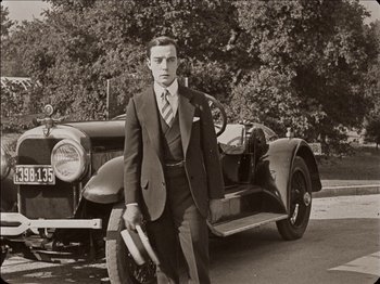 Movie still from “Seven Chances” (1925), directed by Buster Keaton – A man standing next to an old car holding a book; Medium shot, Low angle