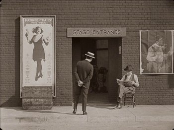 Movie still from “Seven Chances” (1925), directed by Buster Keaton – A man in a suit and a hat standing in front of a building; Wide shot, High angle